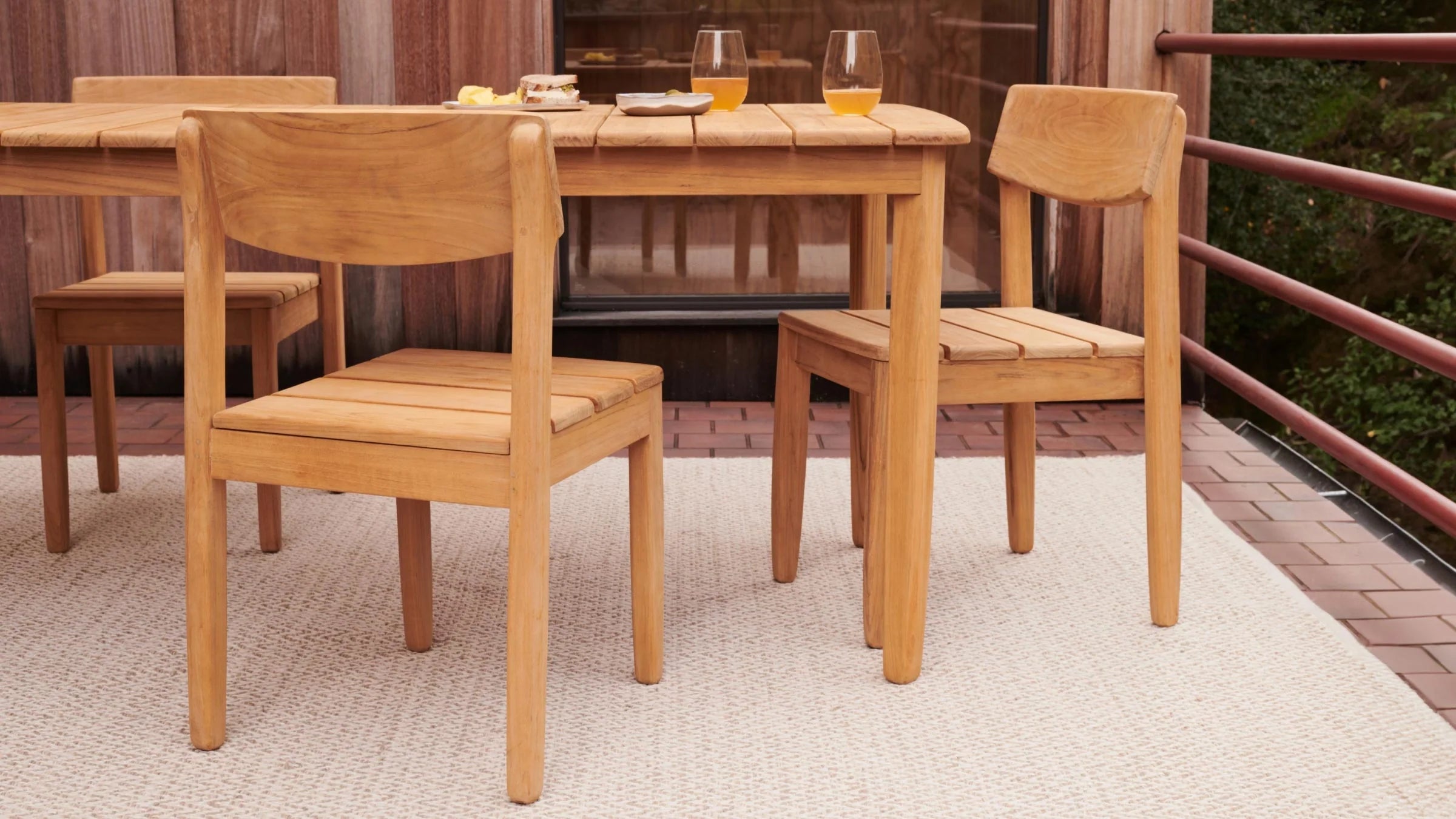 Two light wooden chairs and a matching table set on a beige woven rug on an outdoor patio with a wooden wall background.