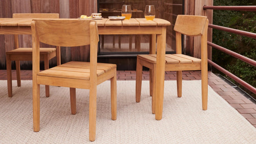 Two light wooden chairs and a matching table set on a beige woven rug on an outdoor patio with a wooden wall background.