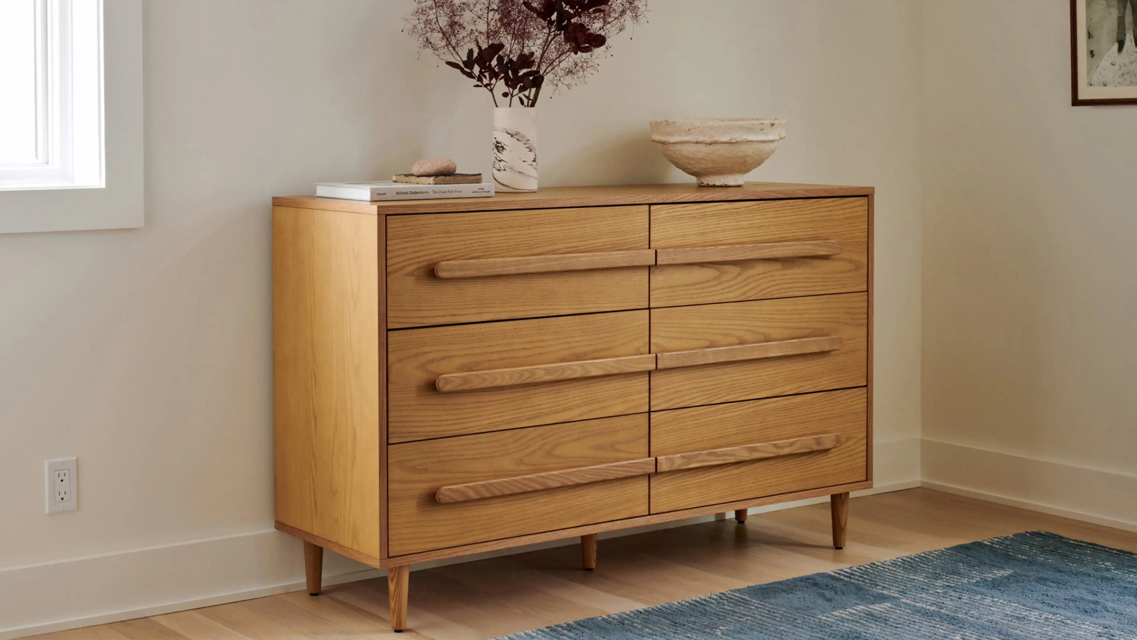 Modern wooden dresser with six drawers and long horizontal handles, decorated with a vase of dried flowers and a bowl.