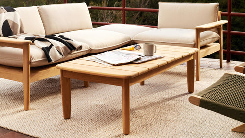 Outdoor wooden coffee table with magazines, mug, and sunglasses on woven rug, accompanied by beige cushioned sofas.