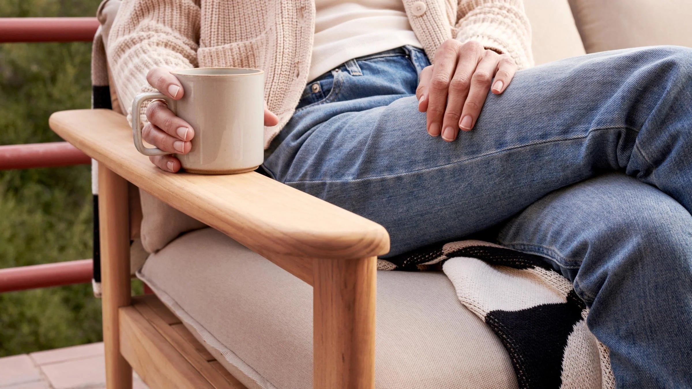 Close-up of a person sitting on a cushioned wooden outdoor chair, holding a beige ceramic mug, wearing jeans and a cream sweater.