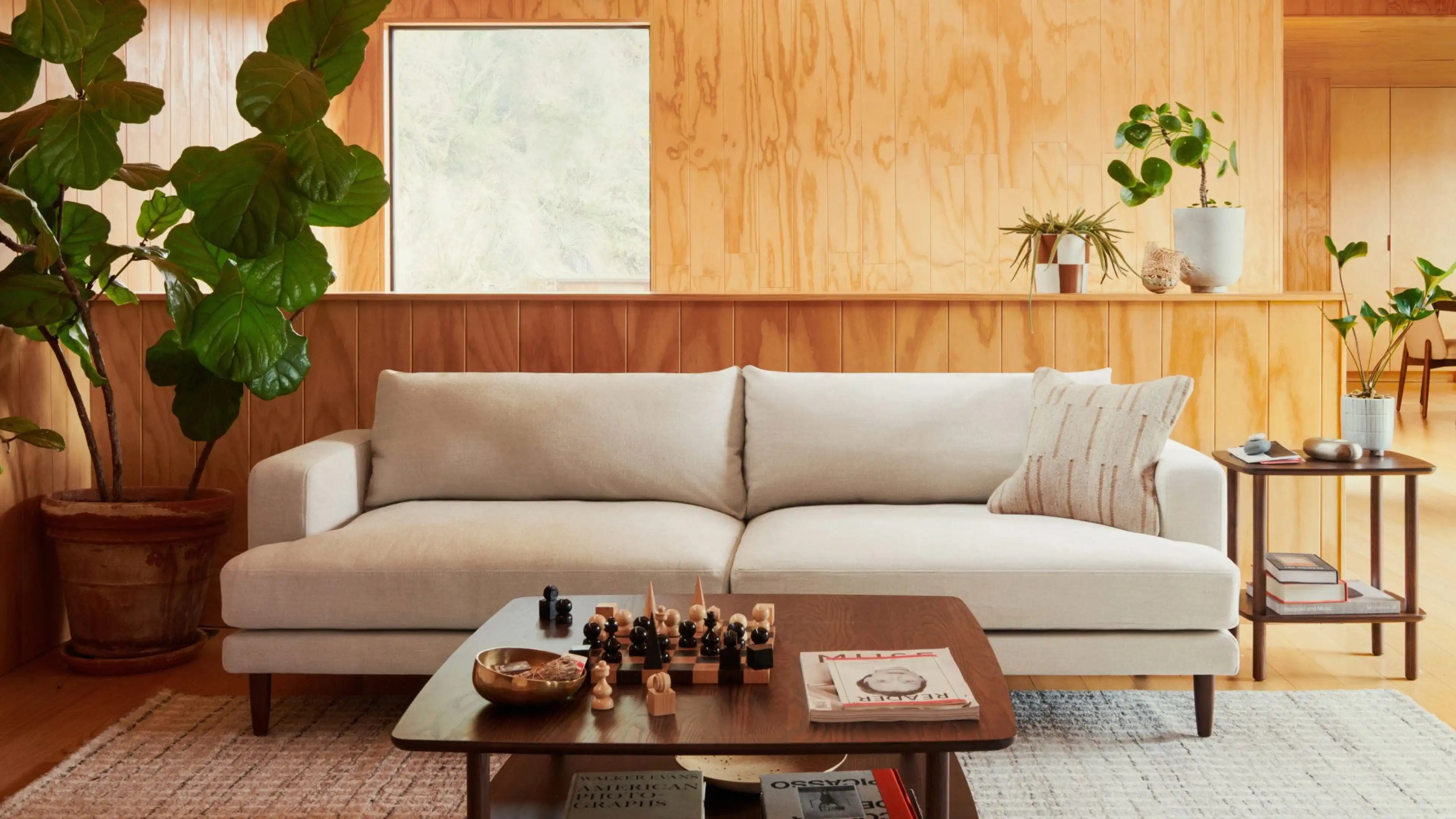 Light beige modern sofa with a textured pillow in a wooden-paneled living room, accompanied by a coffee table and side table.