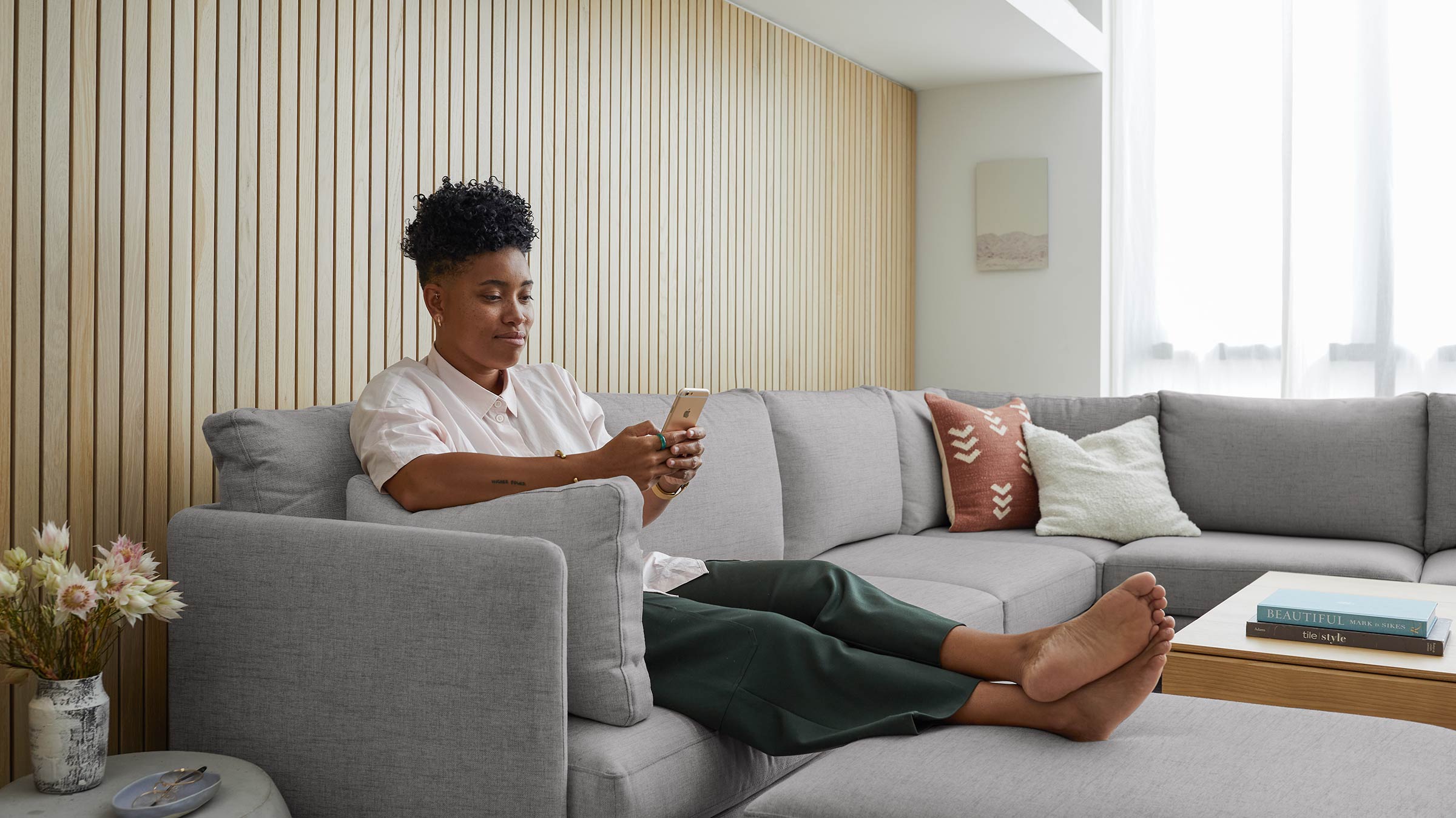 Person sitting barefoot on a light gray sectional sofa with decorative pillows, using a smartphone in a modern living room.