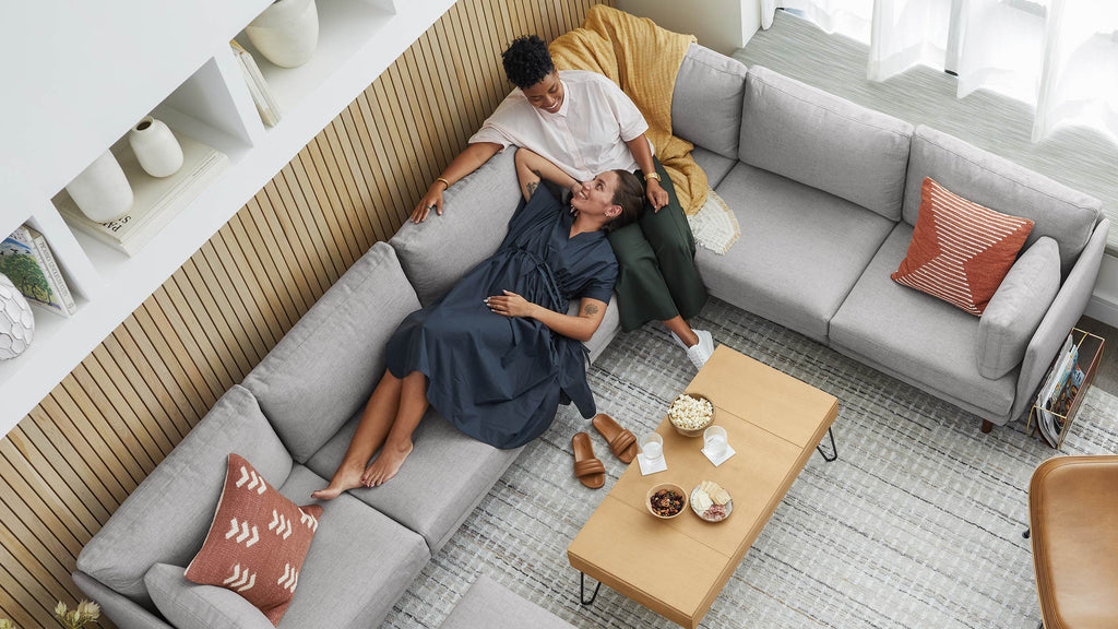 Above view of two women relaxing on a modern light gray sectional sofa with orange patterned pillows in a cozy living room.