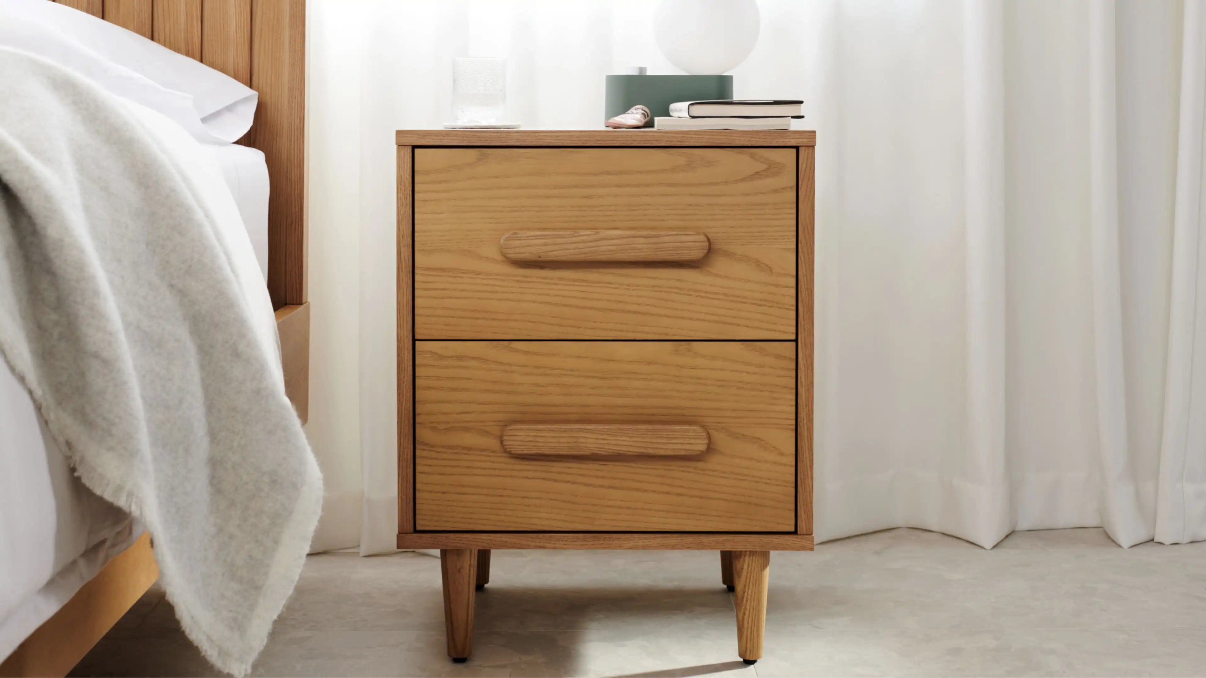 Wooden two-drawer bedside table with rounded handles, standing on four tapered legs, next to a bed with white bedding and soft curtain background.