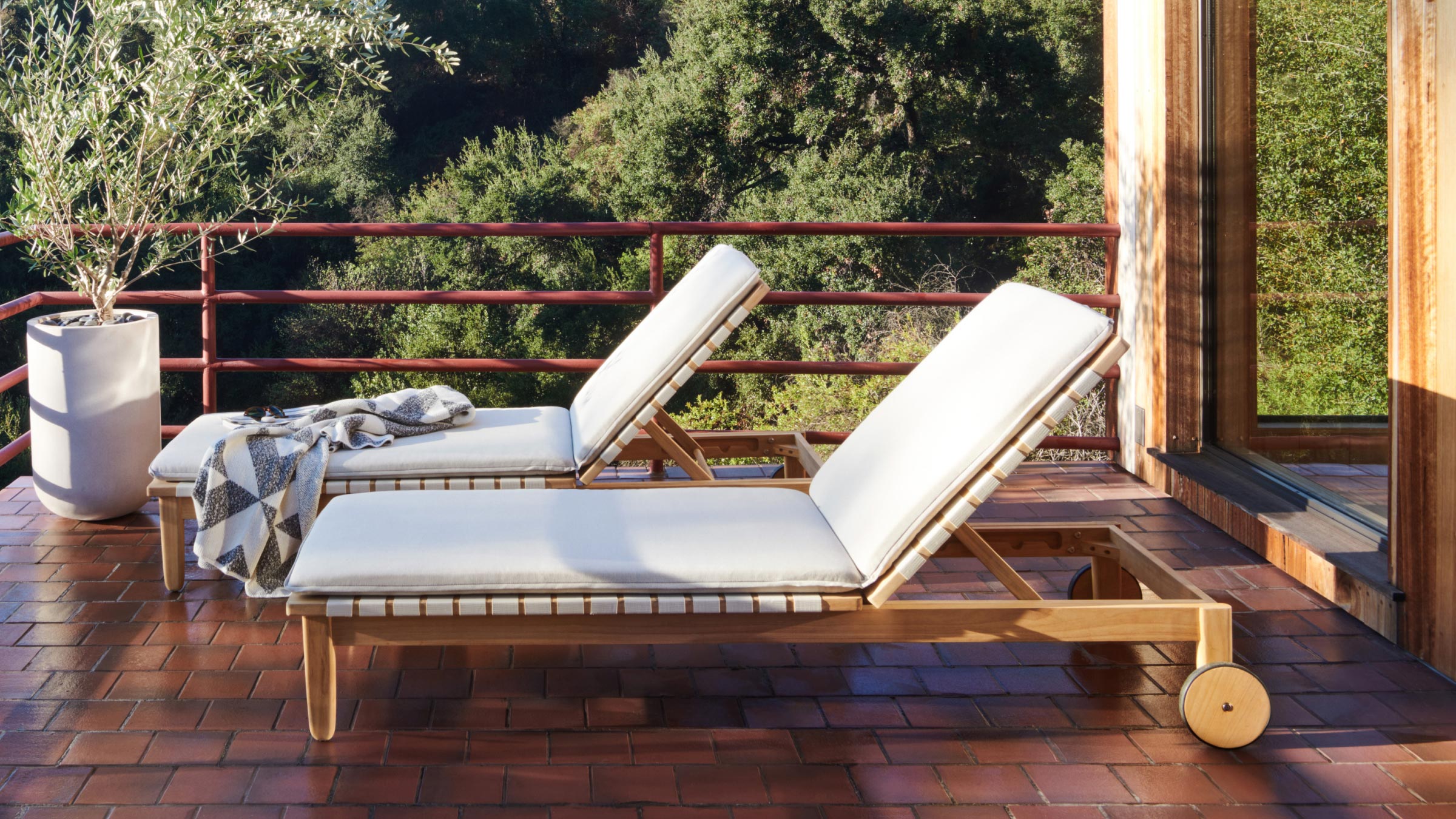 Two wooden outdoor lounge chairs with white cushions on a tiled patio, surrounded by greenery and a potted plant.