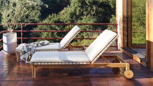 Two wooden outdoor lounge chairs with white cushions on a tiled patio, surrounded by greenery and a potted plant.