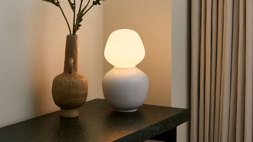 White ceramic table lamp with rounded, modern design and warm light, placed on dark textured surface beside a brown vase with green stems.