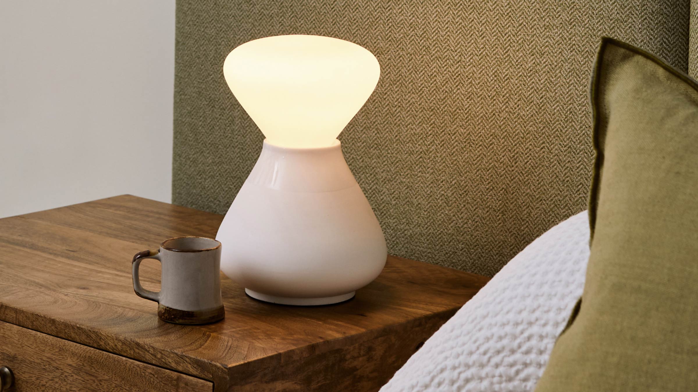 White ceramic table lamp with a rounded base and soft glowing conical diffuser on wooden bedside table.