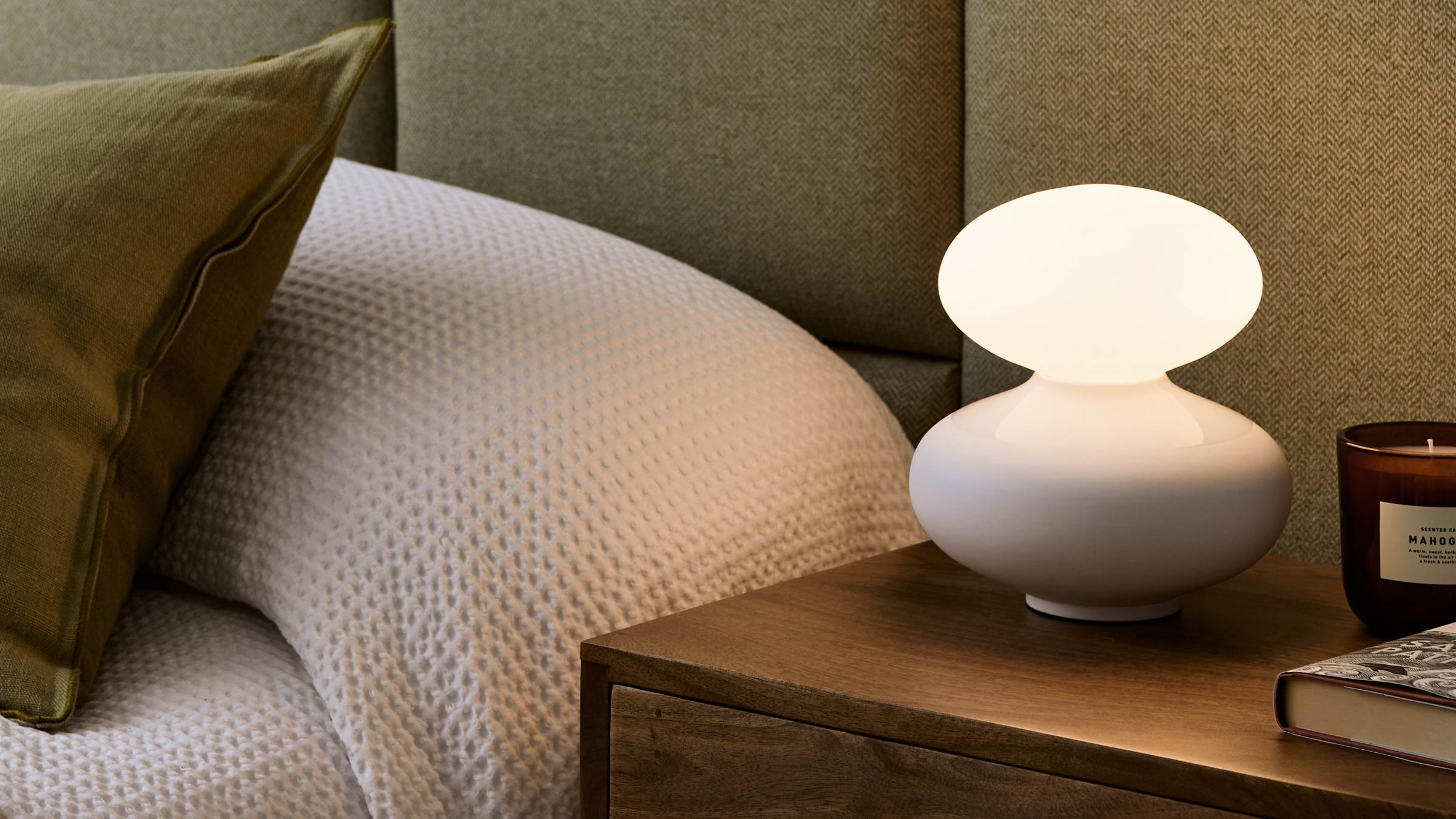 Modern white table lamp with a rounded base and oval shade, placed on a wooden nightstand beside a bed.