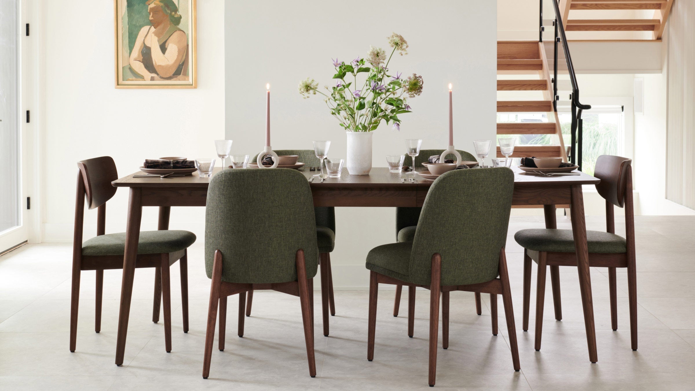 Modern dining table set with six green upholstered chairs, wooden table, decorative candles, and flower vase centerpiece in a bright room.