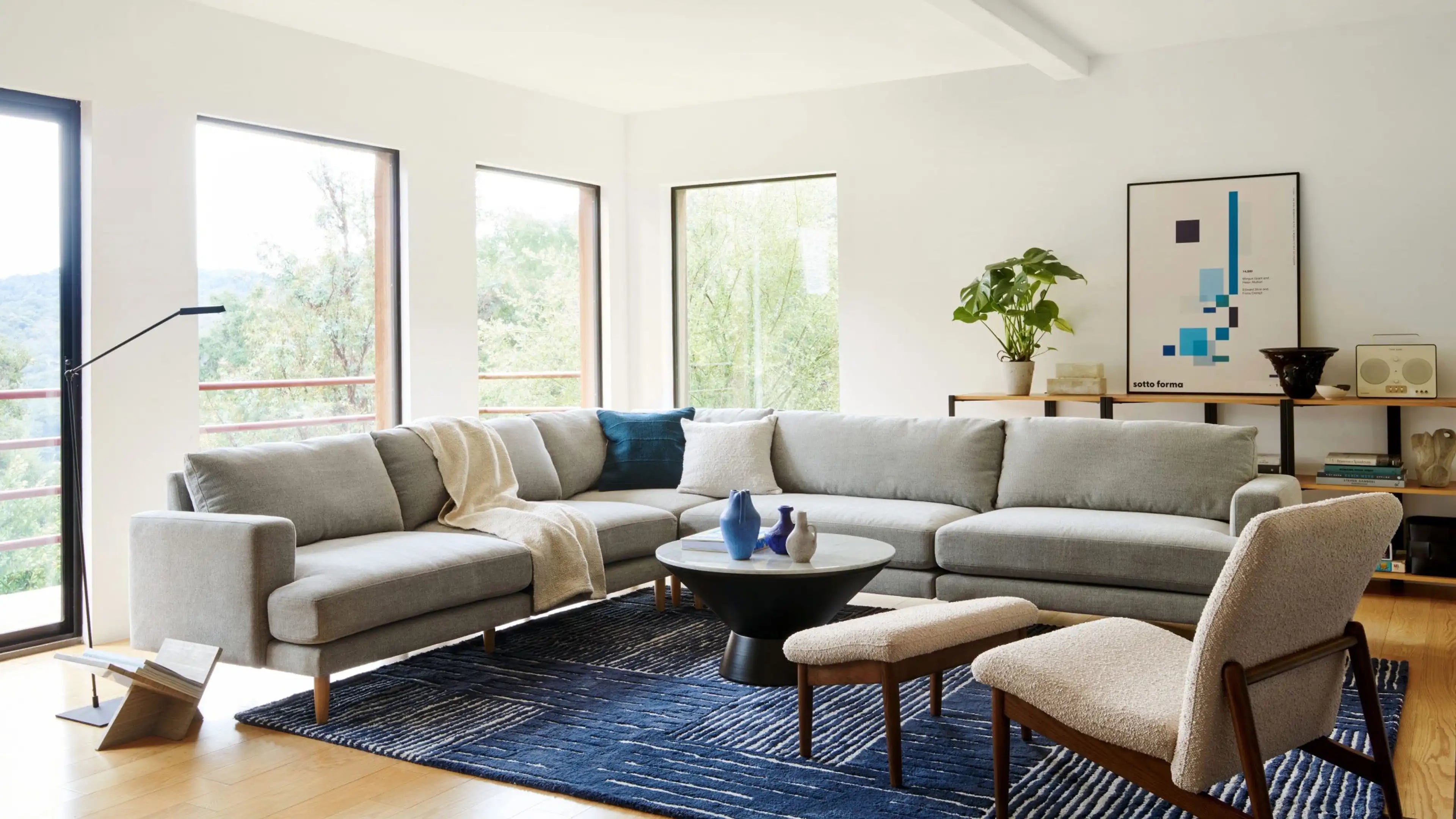 Modern living room with a large light gray sectional sofa, wooden floor, blue patterned rug, round black coffee table, and upholstered armchair with matching bench.