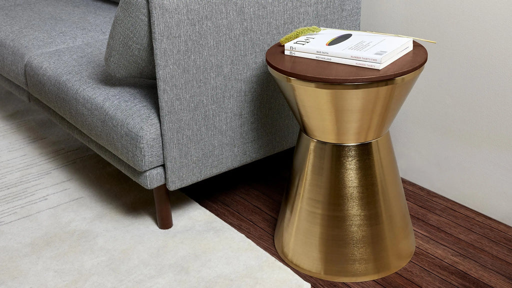 Modern hourglass-shaped metallic gold side table with round wooden top, styled with books and dried flower beside gray fabric sofa.