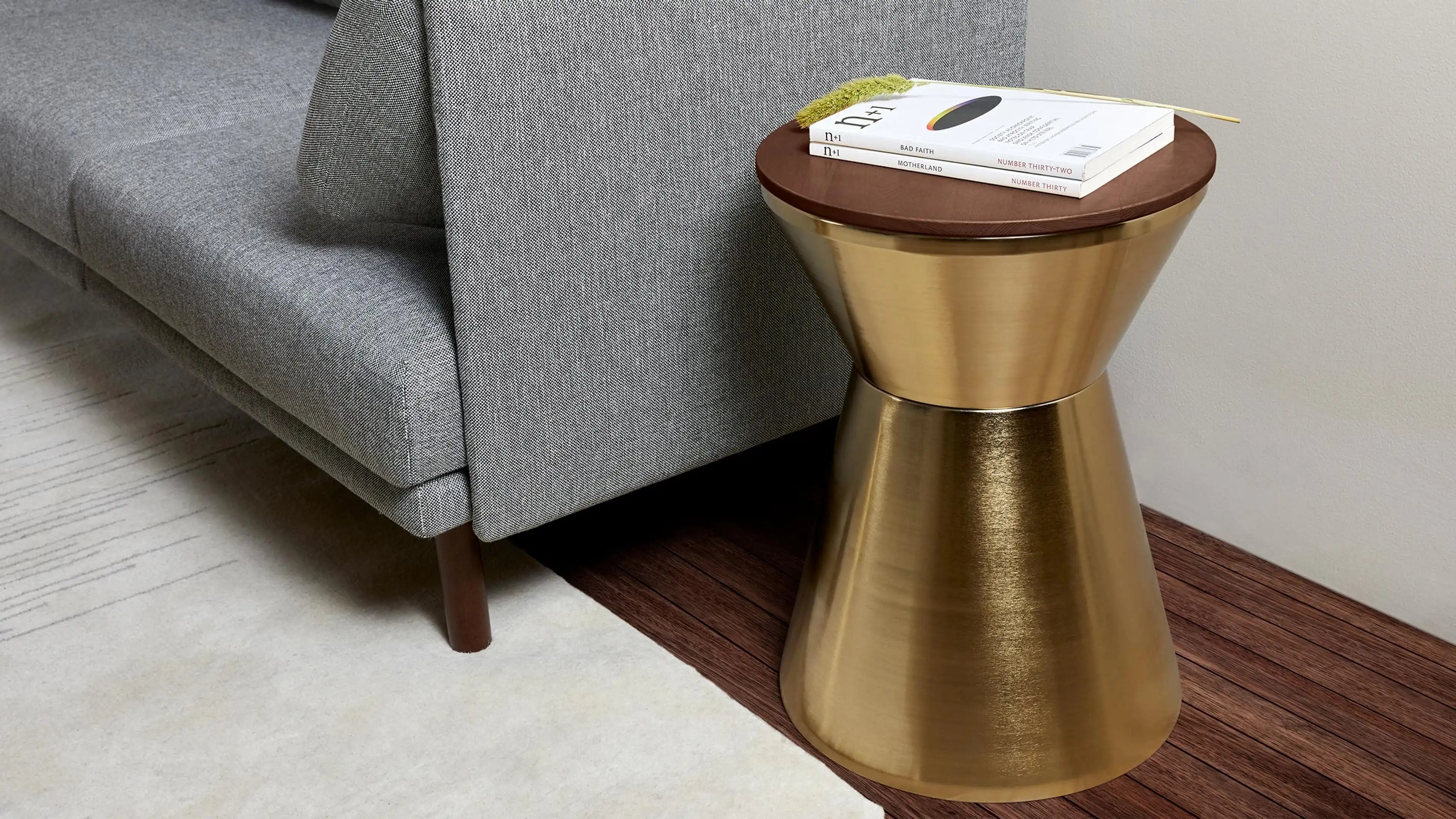 Modern hourglass-shaped metallic gold side table with round wooden top, styled with books and dried flower beside gray fabric sofa.