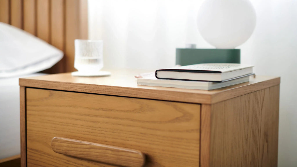 Close-up of a wooden nightstand with a drawer, a stack of books, a glass of water, and a round white lamp on top.