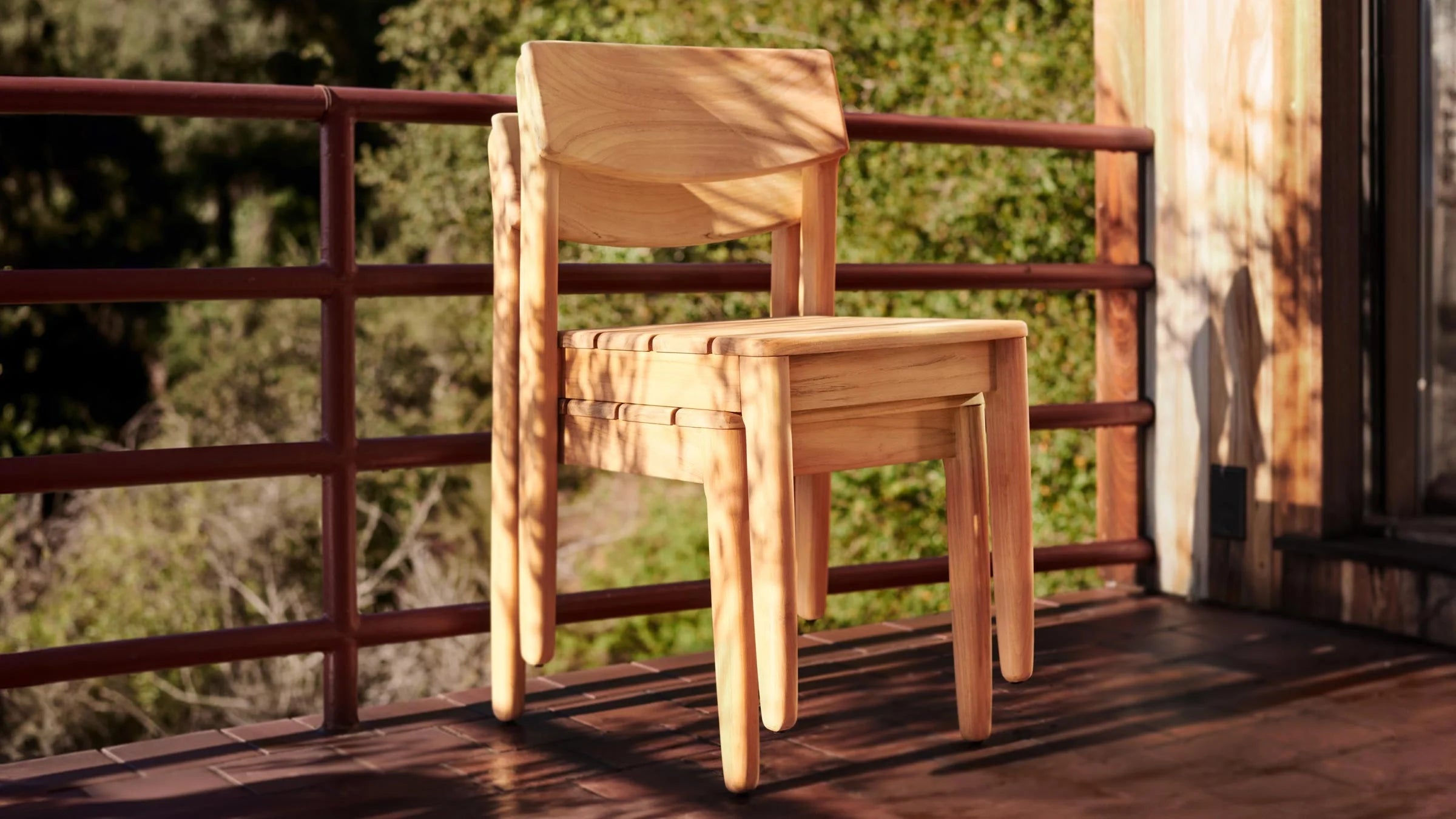 Stacked light wooden chairs with curved backrests, placed on a sunlit outdoor wooden deck near a metal railing.
