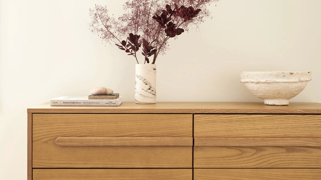 Light wooden dresser with minimalist horizontal handle, topped with a marble vase holding dried branches, stacked books, and a rustic white bowl.