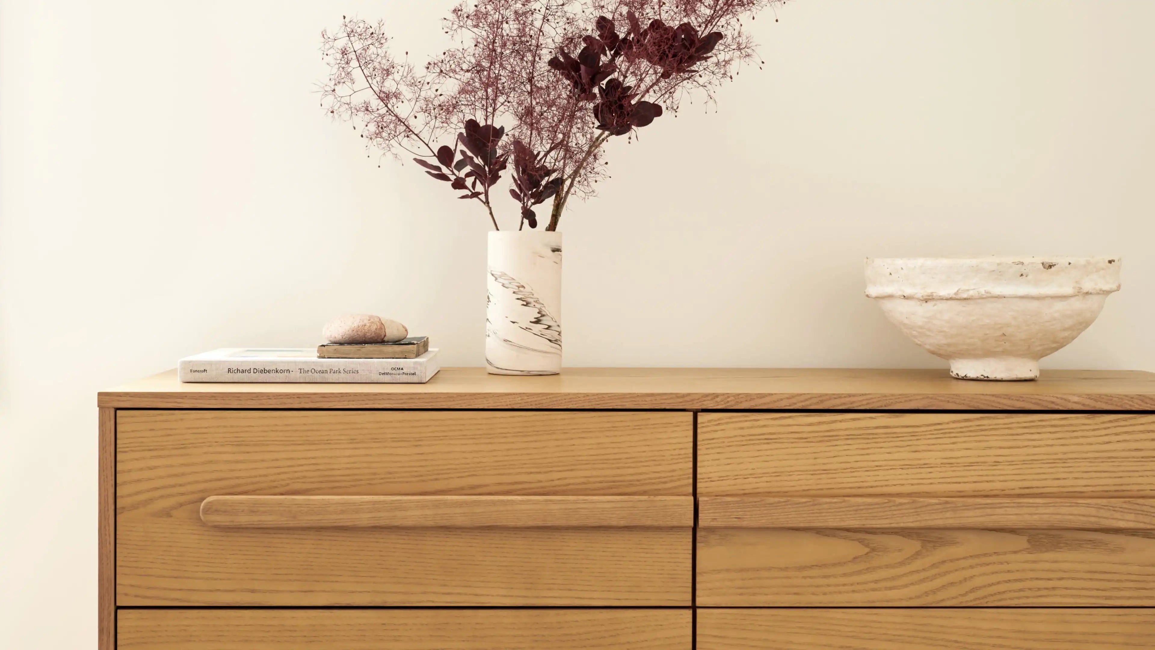 Light wooden dresser with minimalist horizontal handle, topped with a marble vase holding dried branches, stacked books, and a rustic white bowl.