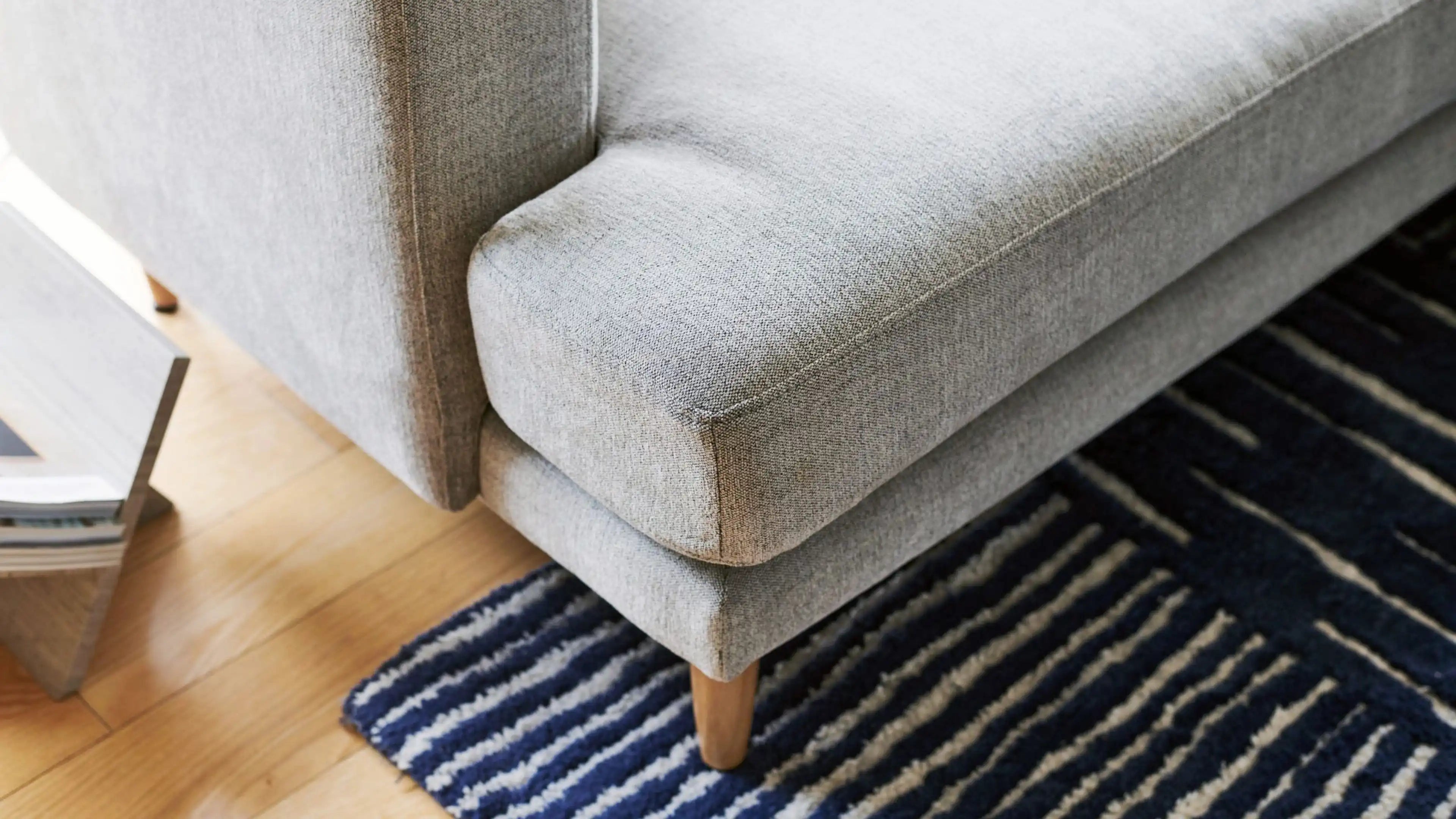 Close-up of a modern grey fabric sofa corner with wooden legs on a striped blue and white rug over light hardwood flooring.