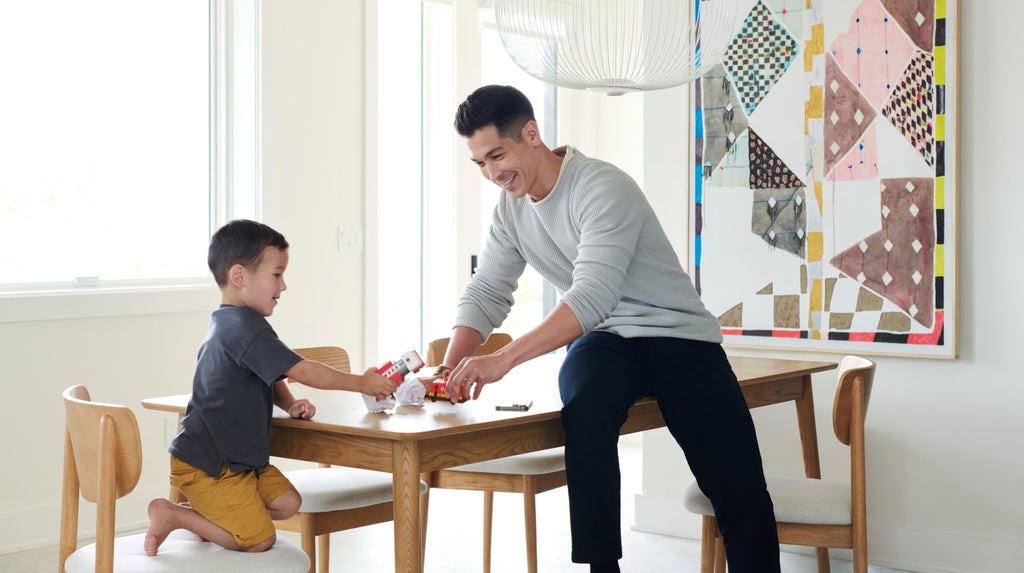 A man and a young boy playing with toy vehicles on a wooden dining table in a bright, modern room.