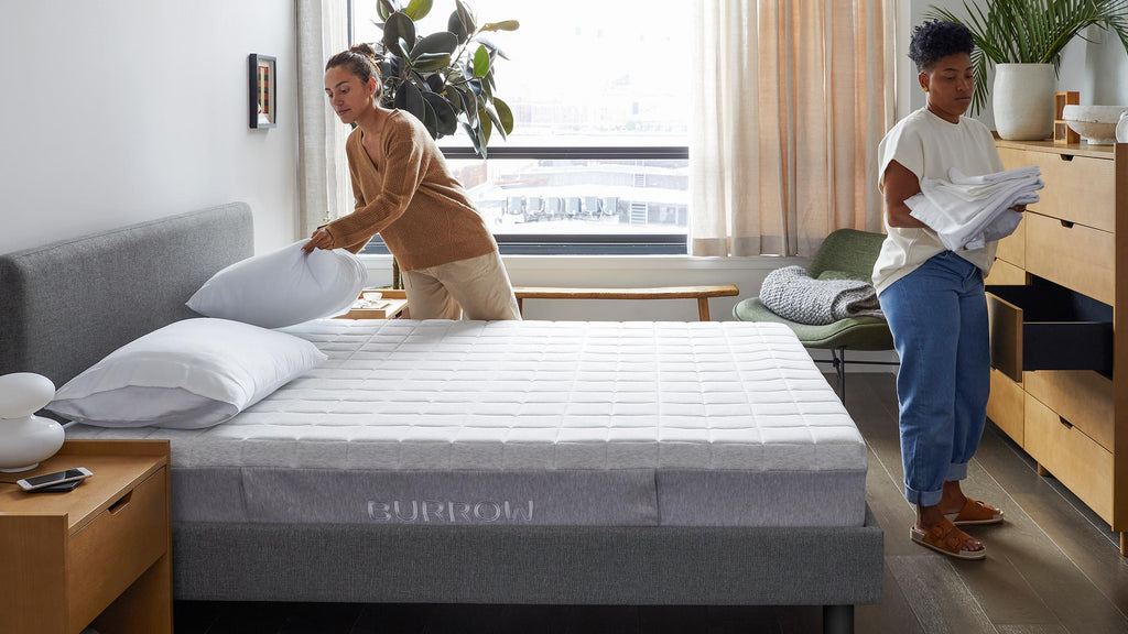 Two people making a bed with a gray Burrow mattress in a modern, bright bedroom with wooden furniture.