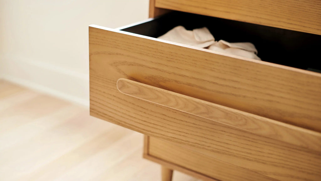 Light wood drawer partially opened, revealing folded white clothes inside, with smooth horizontal handle detail.