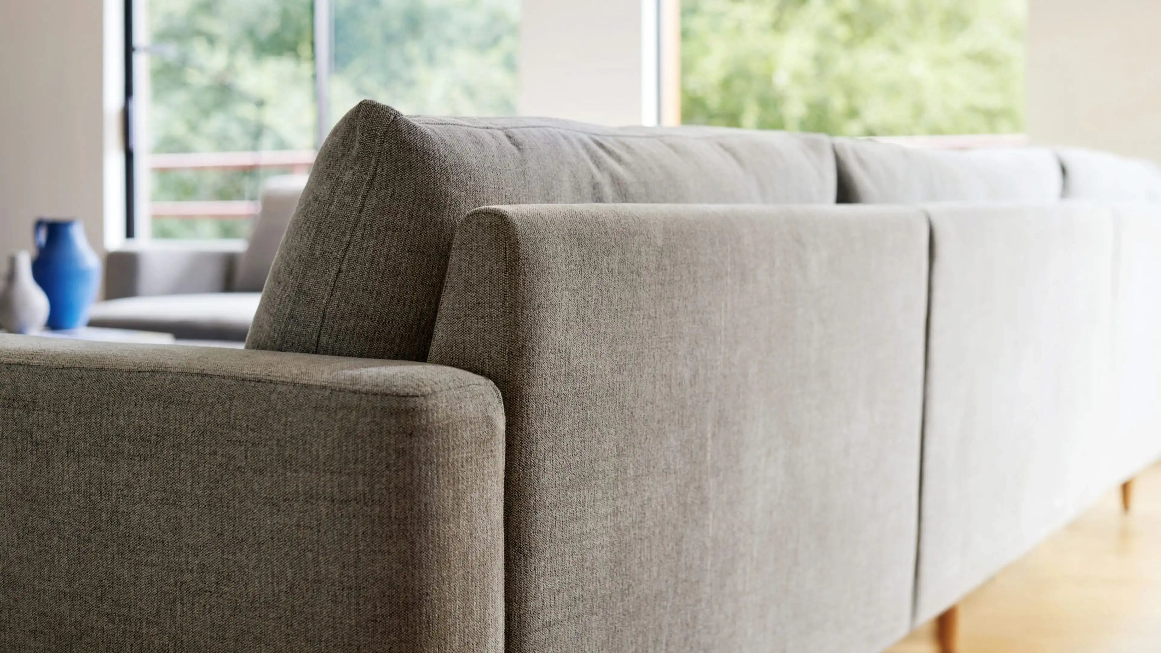 Close-up side view of a modern, light gray fabric sofa with clean lines and wooden legs in a bright living room.