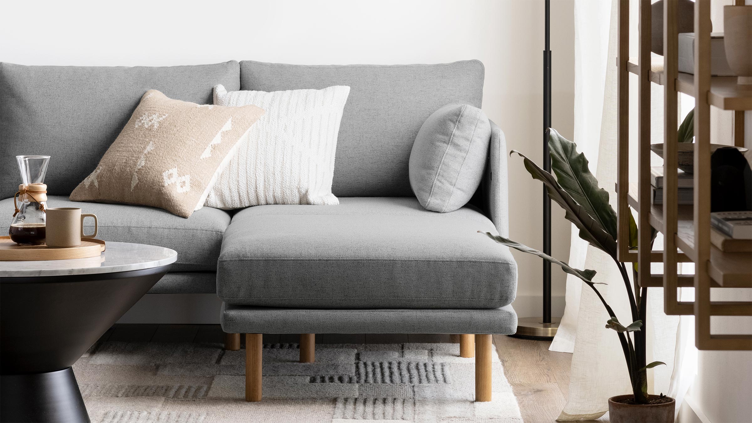 Modern light gray sectional sofa with three decorative pillows, wooden legs, next to a black coffee table and potted plant.
