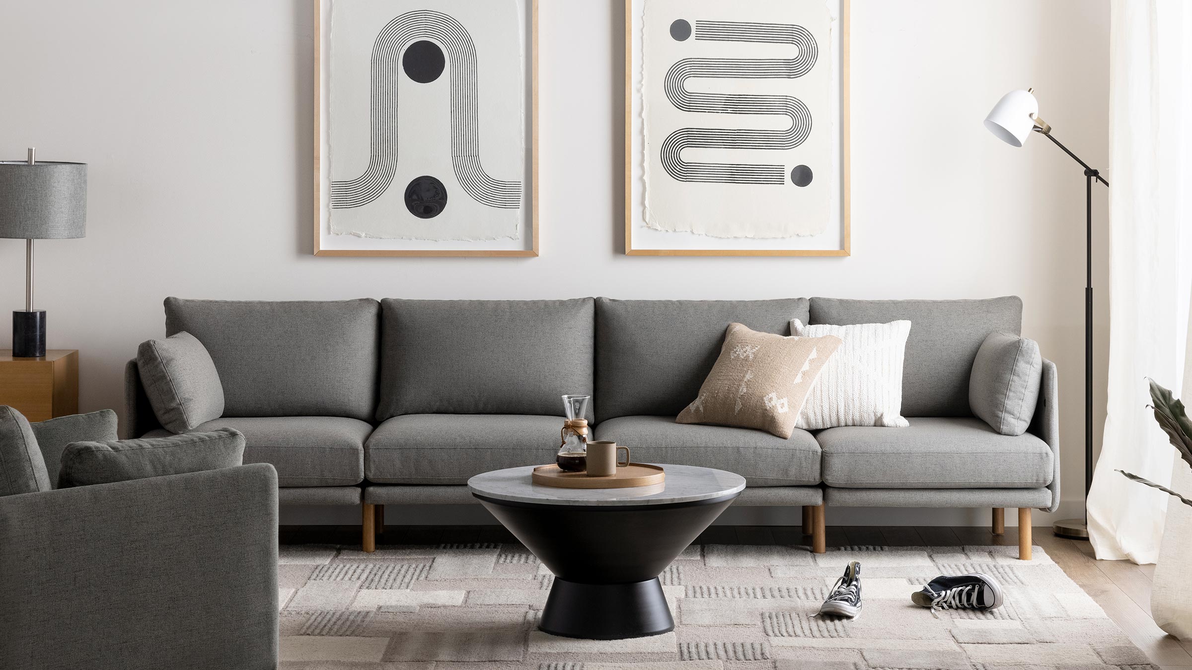 Modern living room with a long gray sectional sofa, round black coffee table, abstract wall art, floor lamp, and cozy neutral-toned decor.