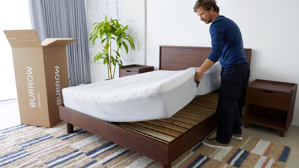 Man placing a gray Burrow mattress onto a wooden bed frame in a modern, minimal bedroom with a plant and nightstands.