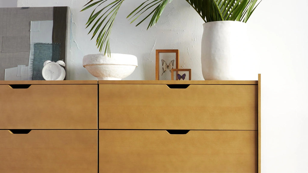 Light wood dresser with four drawers, decorated with a white alarm clock, white bowl, framed butterfly art, and a large white vase with green palm leaves.