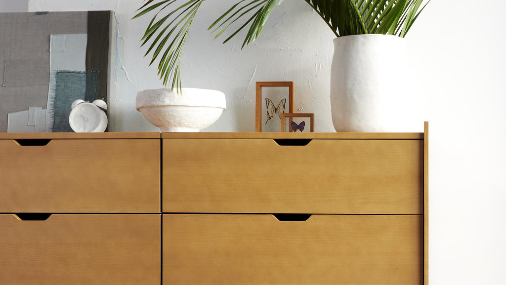 Light wood dresser with four drawers, decorated with white ceramic pot, bowl, framed butterflies, and white alarm clock.