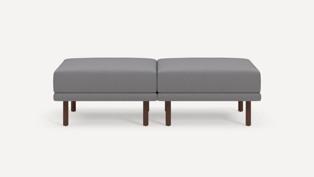 Modern gray fabric ottoman bench with two cushioned seats and dark wooden legs, featuring a sleek and minimalist design.