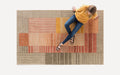 Top-down view of a person sitting on a modern area rug with geometric patterns in earthy tones of beige, orange, and brown.