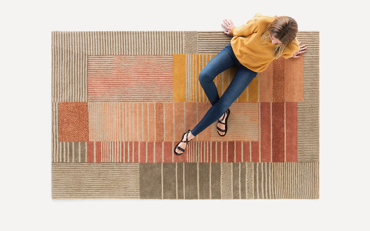 Top-down view of a person sitting on a modern area rug with geometric patterns in earthy tones of beige, orange, and brown.