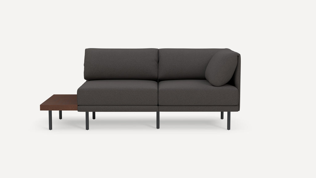 Modern dark gray two-seater sofa with cushioned backrest, a round pillow on one side, and an attached wooden side table with metal legs.