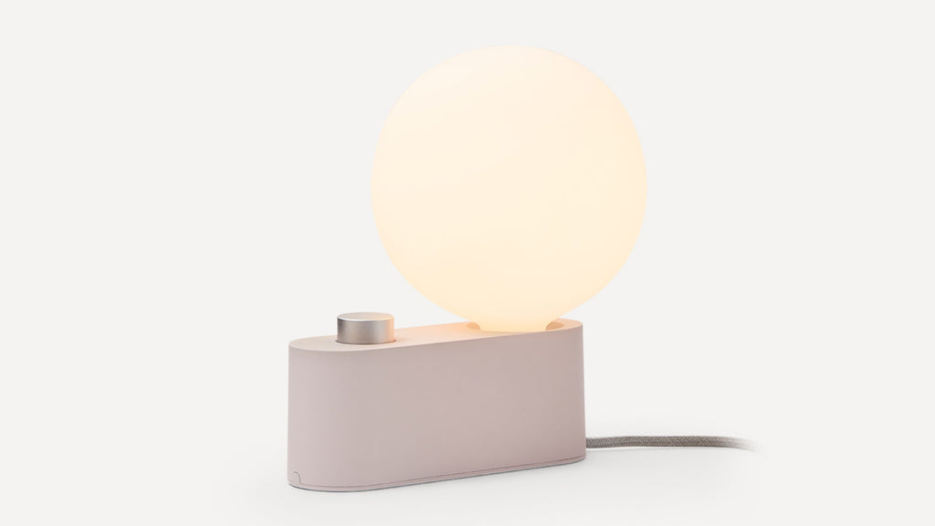 Modern table lamp with a glowing round white bulb, matte beige base, and a silver knob, on a white background.