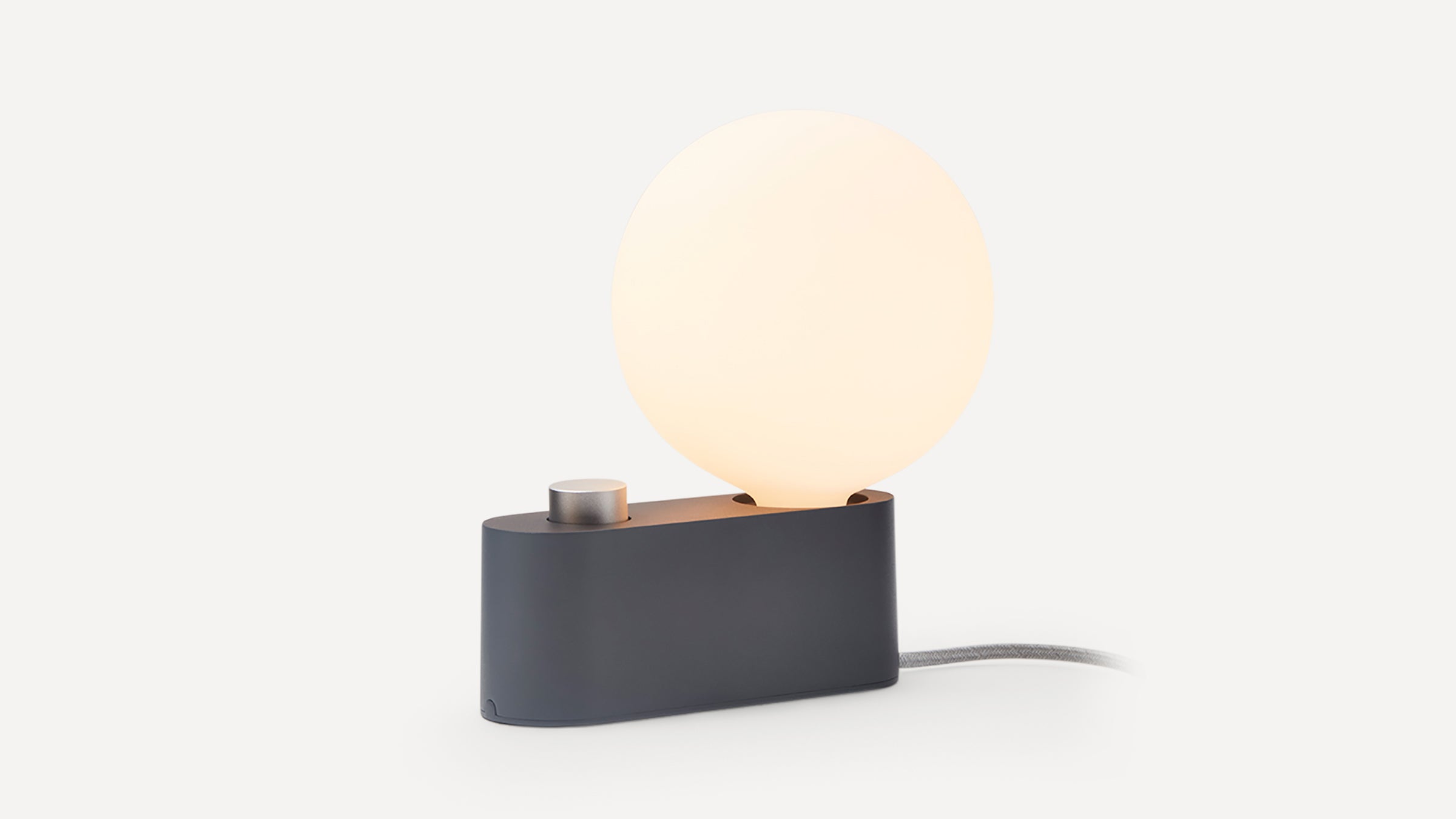 Minimalist black desk lamp with a round white glowing bulb and a silver control knob on the base, connected by a gray fabric cord.
