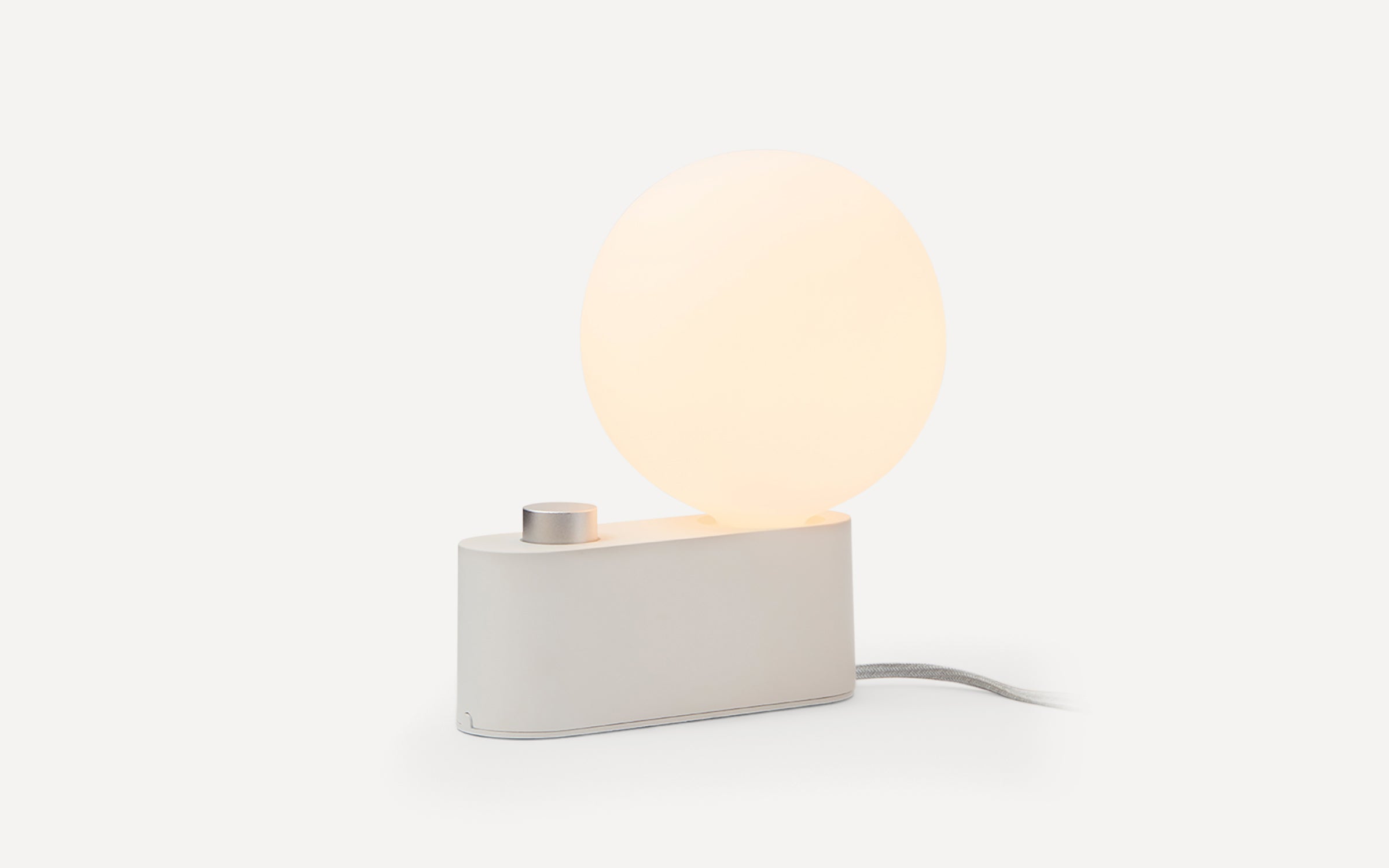 Minimalist white table lamp with a glowing spherical light bulb, featuring a cylindrical base and a fabric-covered power cord.