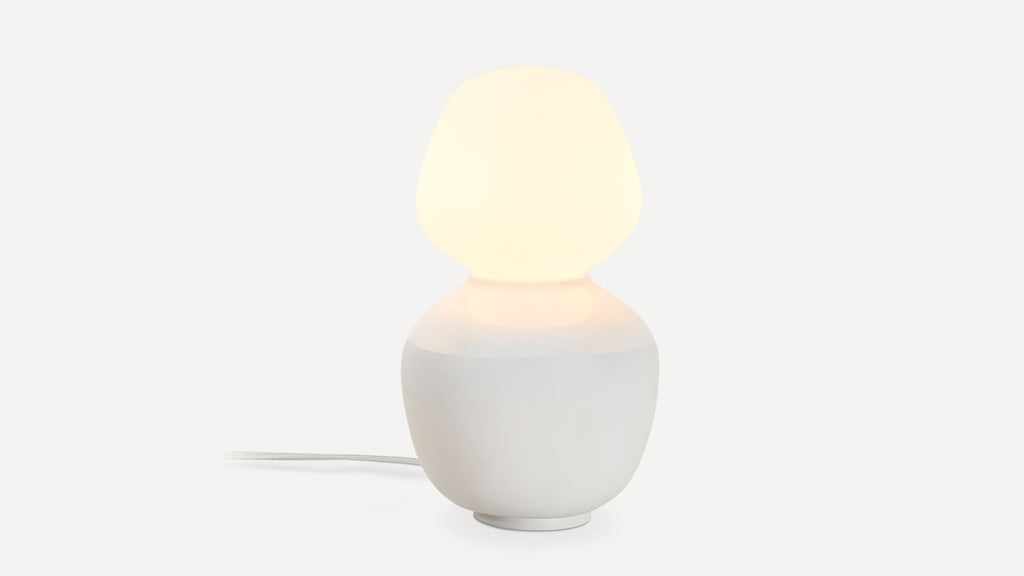 Modern table lamp with a rounded white ceramic base and a soft glowing frosted glass shade, emitting warm light.