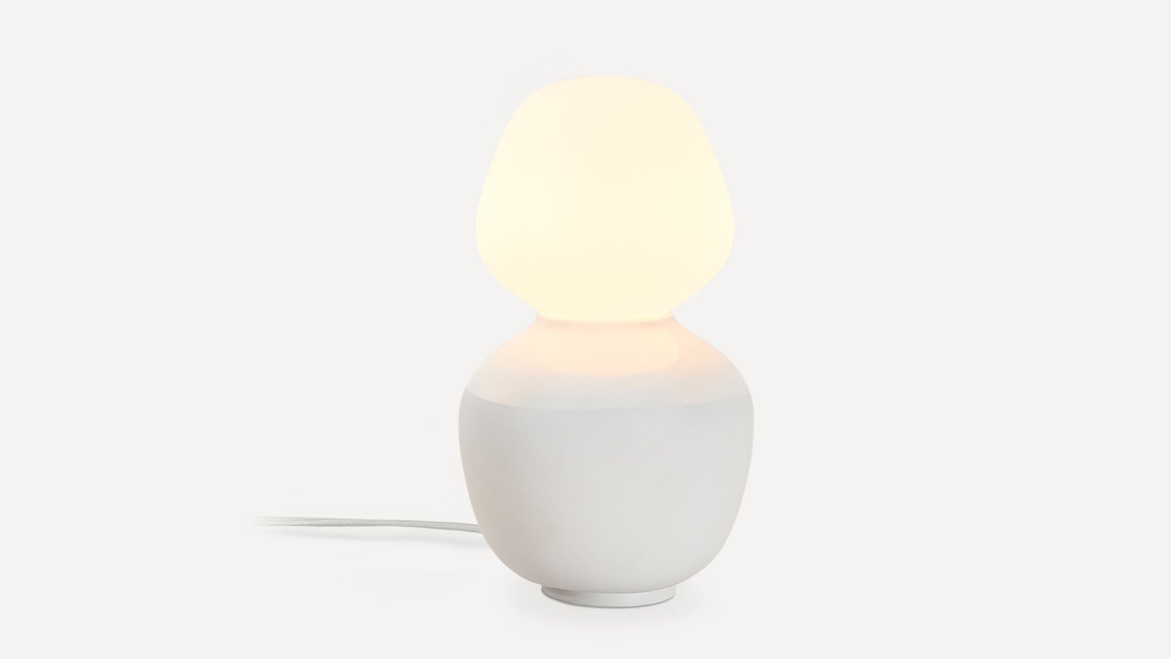 Modern table lamp with a rounded white ceramic base and a soft glowing frosted glass shade, emitting warm light.