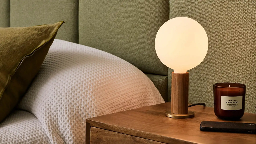 Bedside wooden table lamp with round frosted glass shade, placed next to a brown candle and remote on wooden nightstand.