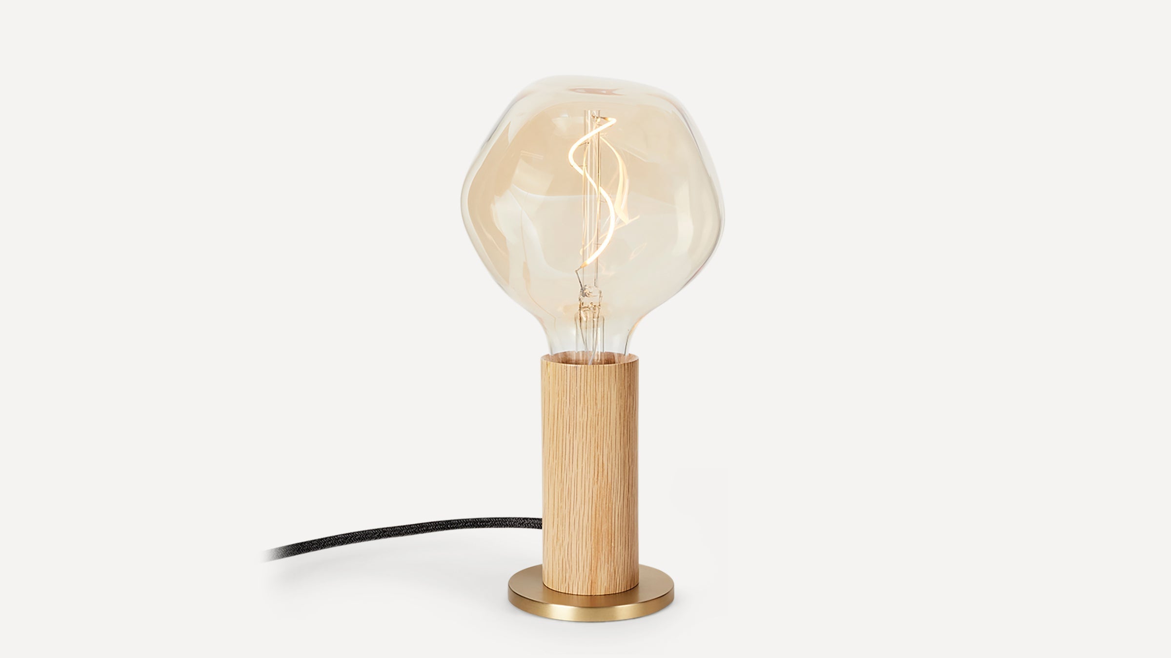 Minimalist table lamp with a wooden cylindrical base, brass circular stand, and a large clear, irregular-shaped light bulb.