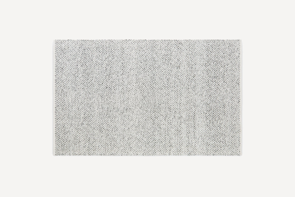 Rectangular rug with a subtle, light gray and white herringbone pattern, featuring neatly finished edges on a plain background.