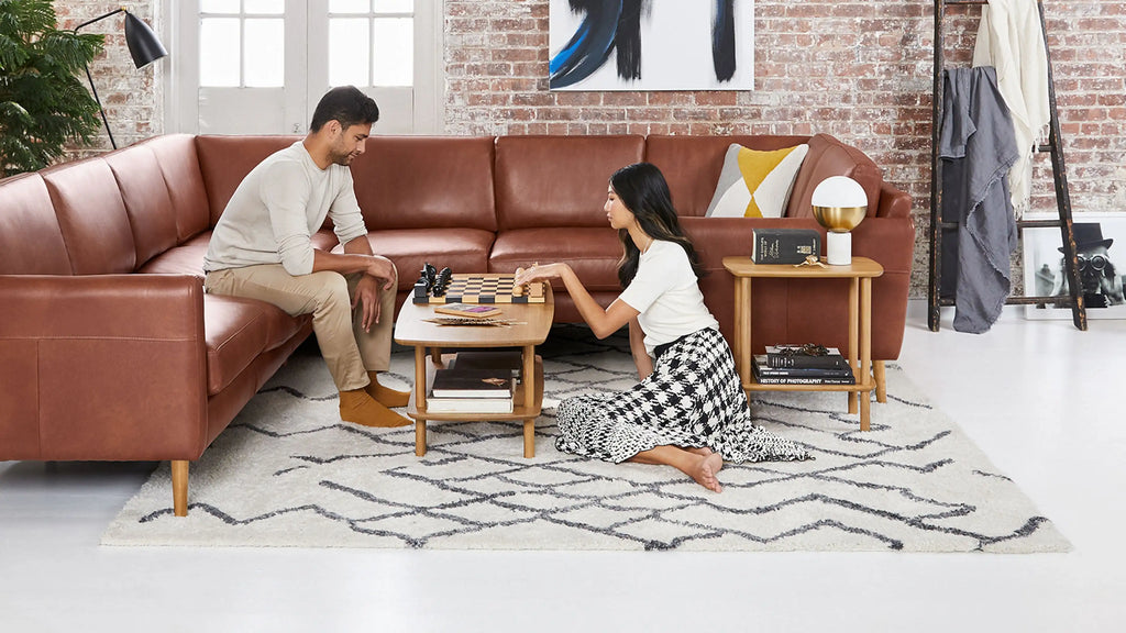 a man and woman sitting on a brown leather sectional sofa in a living room with a white rug and a wooden coffee table