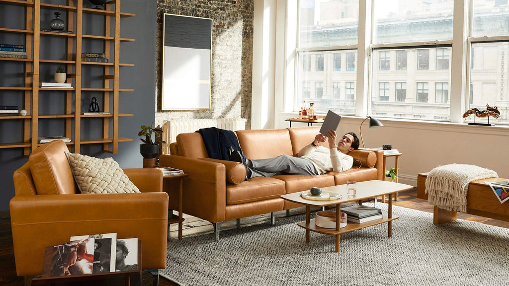 Modern living room with tan leather sofa and armchair, wooden coffee table, large windows, and a man lounging while reading.