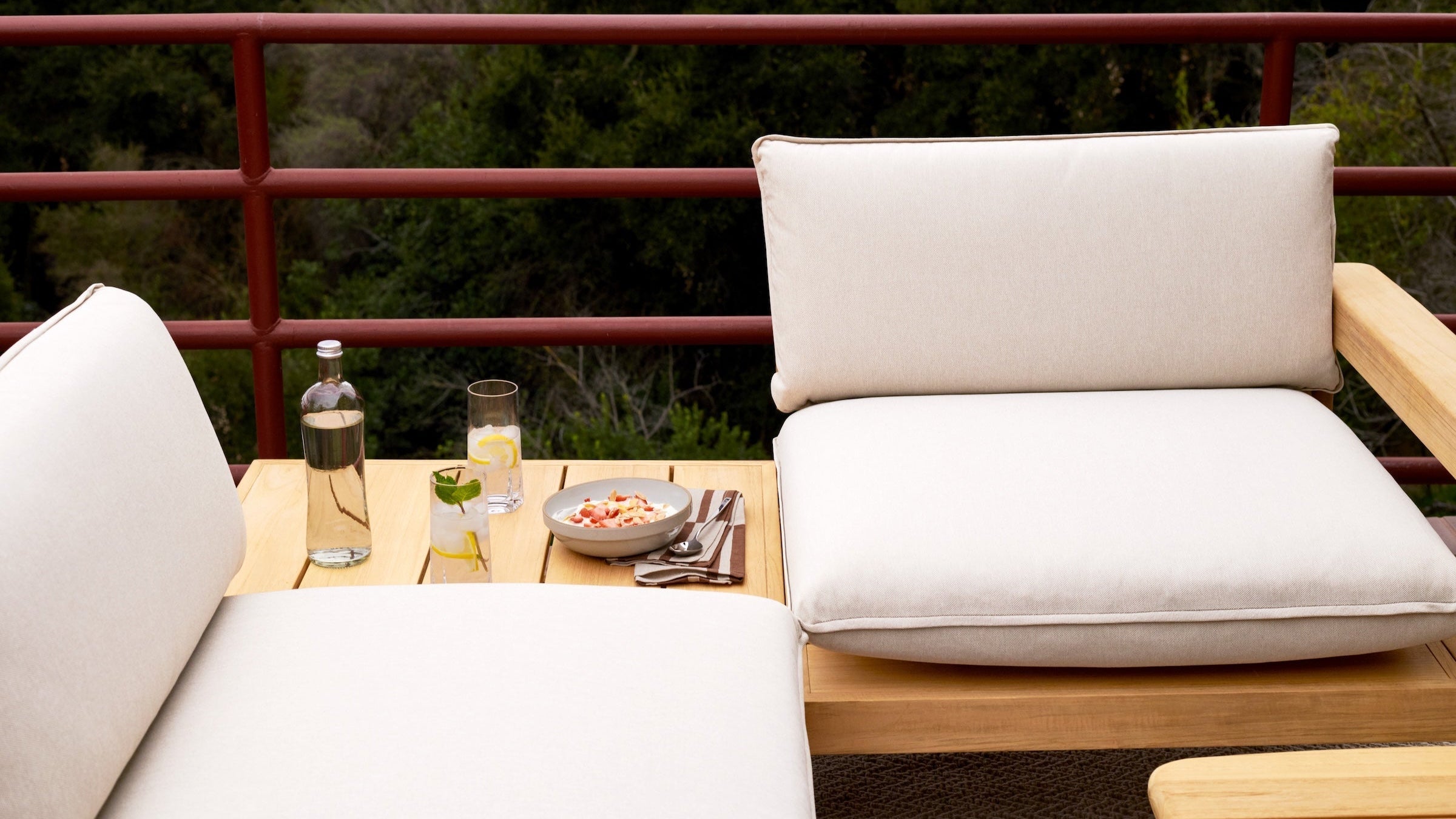 Outdoor wooden sectional sofa set with cream cushions, small wooden table holding water bottle, glasses with lemon and mint, and a bowl with food.