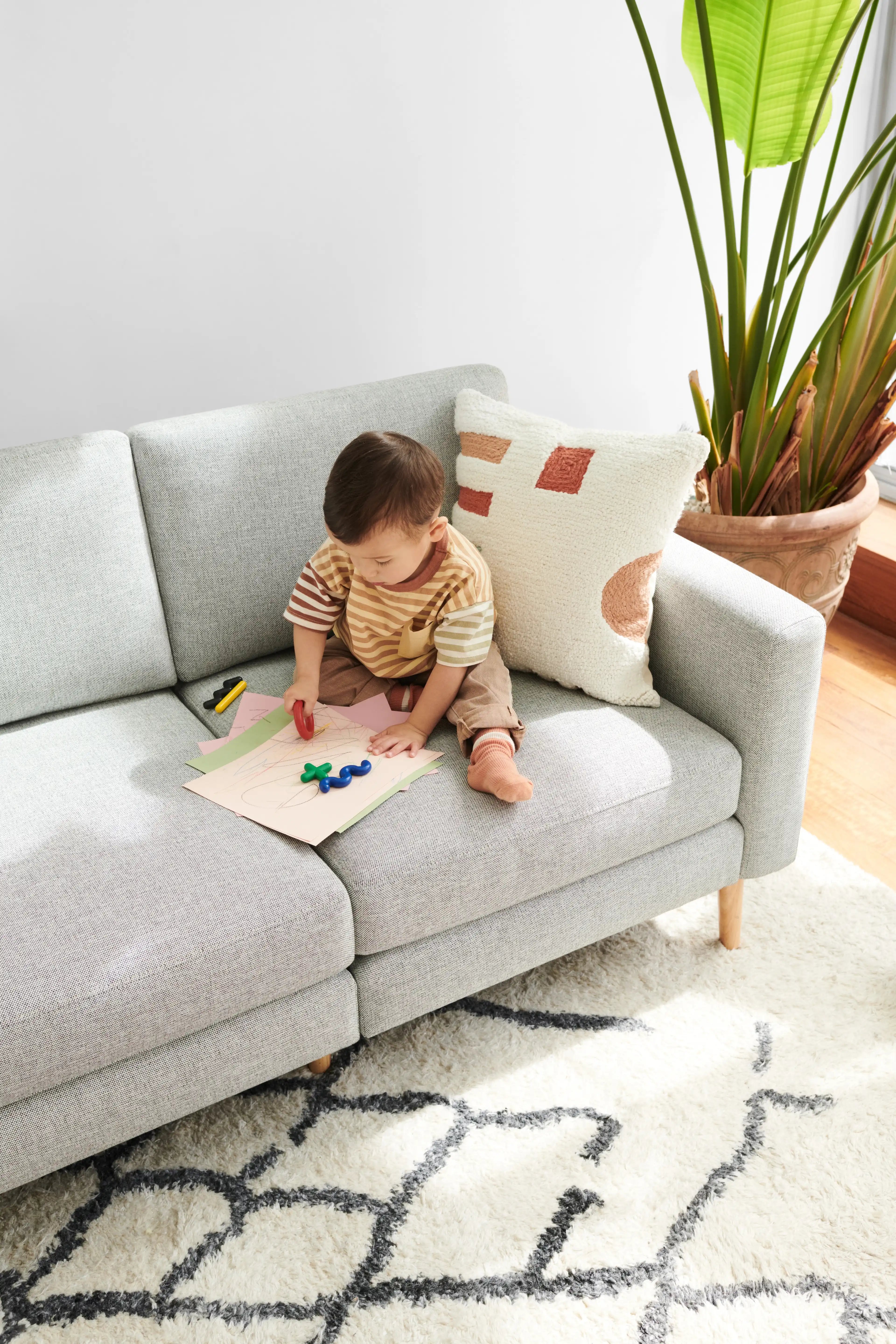 a baby boy sitting on a gray couch with a book and pencils on it