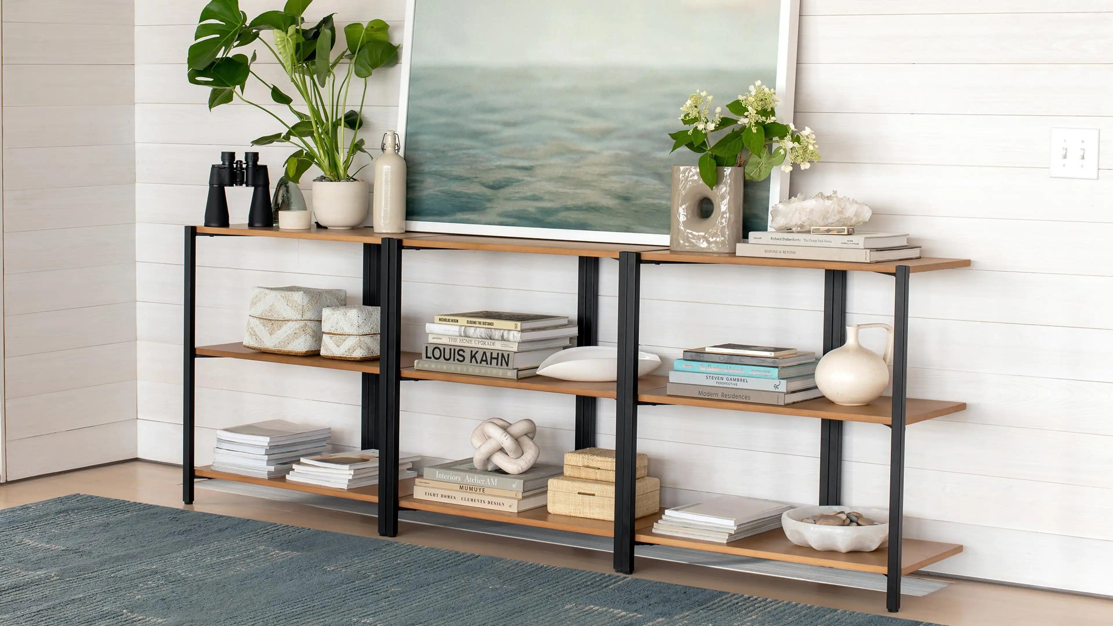 Three-tier wooden shelving unit with black metal frame, decorated with plants, books, vases, and decorative objects against a white shiplap wall.