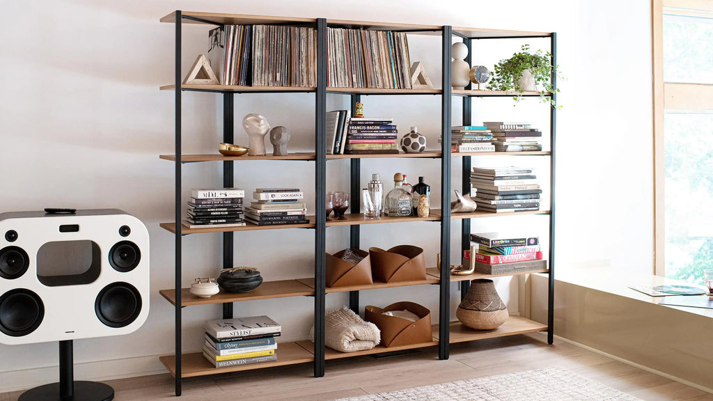 Open black metal frame and wood shelving unit filled with books, decor pieces, plants, and storage baskets in a bright living room.